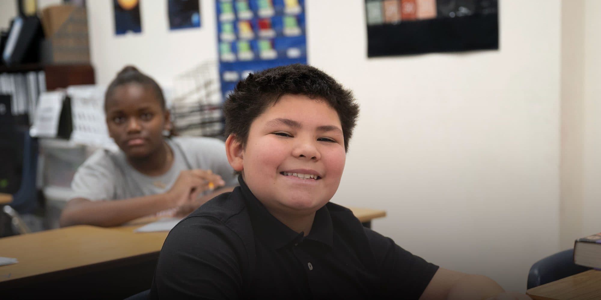 Smiling student in a black polo shirt at a classroom desk, with a classmate working in the background.
