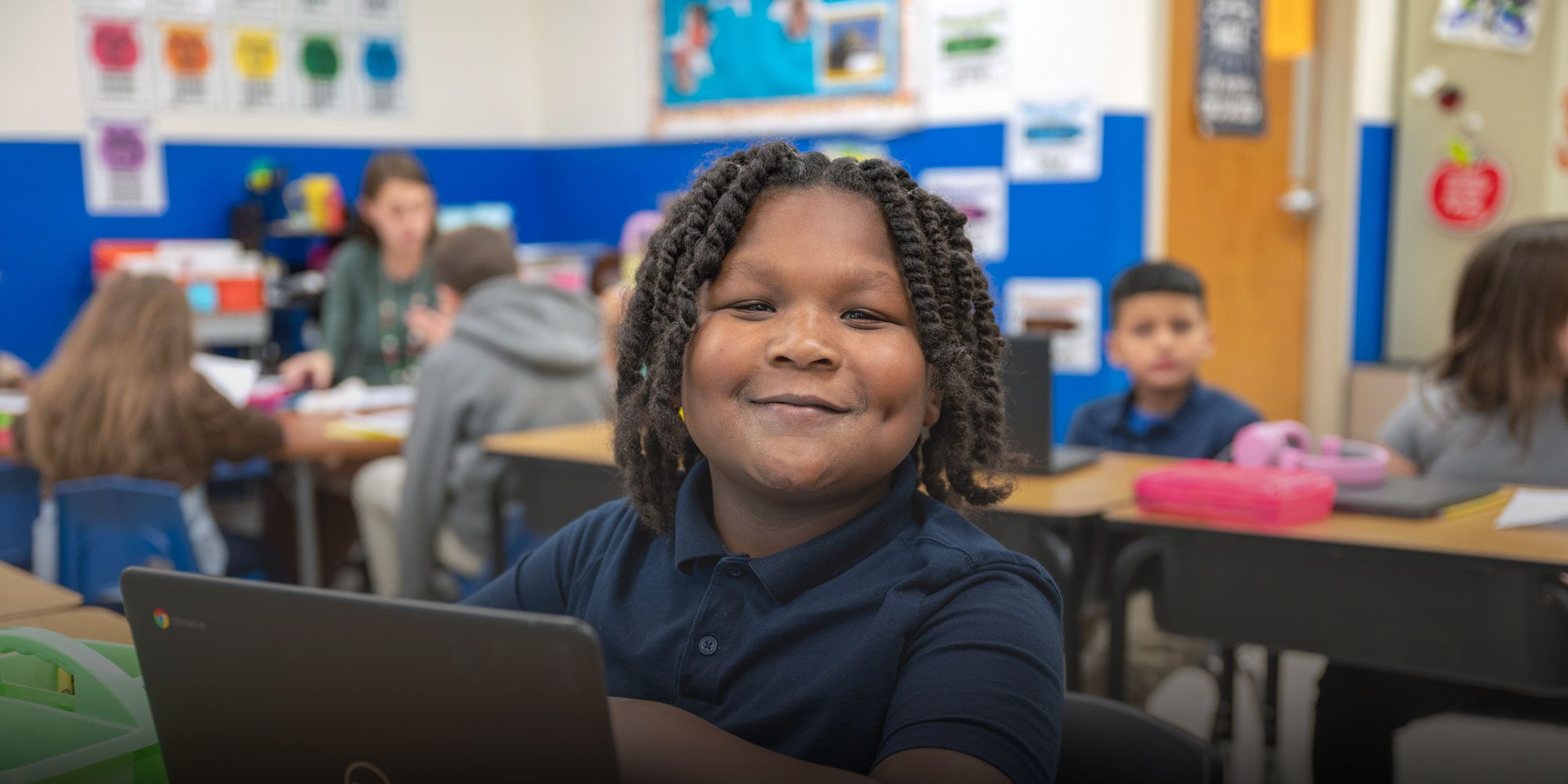 Smiling student with a Chromebook at a desk in a classroom.